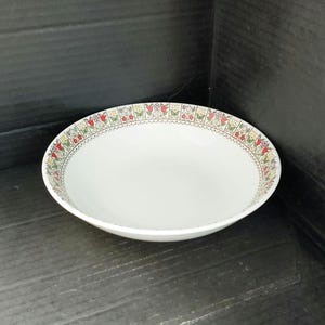 May include: A white ceramic bowl with a decorative floral border featuring red, yellow, and green flowers. The bowl has a wide, shallow design, suitable for serving soups or salads. The bowl is set against a dark background.