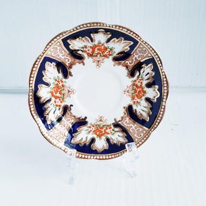 May include: A decorative saucer with a white center and a navy blue and gold rim. The saucer features a floral design in orange and white. The edge of the saucer has a scalloped design.