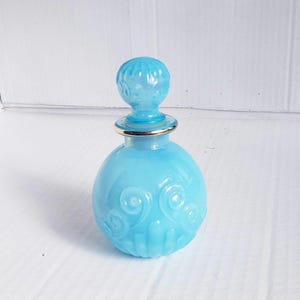 May include: A light blue glass perfume bottle with a spherical body and a matching round stopper. The bottle features embossed swirl patterns and vertical fluting at the base, with a metallic gold-toned band encircling the neck of the vessel.