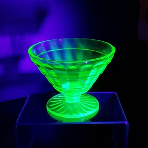 May include: A glowing green glass dessert dish with a pedestal base. The dish has a flared bowl with vertical and horizontal etched lines. The base has a sunburst design. The dish is displayed on a clear acrylic stand.
