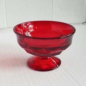 May include: A ruby red glass dessert dish with a pedestal base. The bowl has a rounded shape with a slightly flared rim. The glass has a smooth, glossy finish, and the base is a solid, rounded shape. The dish is likely vintage.