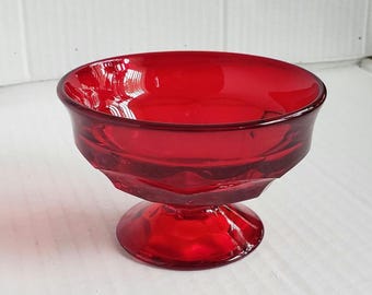 Peut inclure: Un petit plat à dessert en verre rouge rubis avec un pied. Le bol a une forme arrondie avec un bord légèrement évasé. Le verre a une finition lisse et brillante, et la base est de forme arrondie. Le plat est probablement vintage.