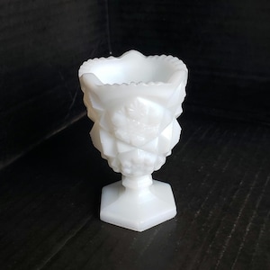 May include: A white milk glass toothpick holder featuring a pressed diamond pattern and a scalloped rim. The small vessel stands on a hexagonal base and displays a raised floral motif on the exterior surface, showcasing a translucent quality.
