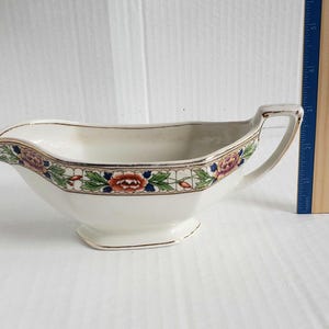 May include: Cream-colored porcelain gravy boat with a floral border in shades of red, blue, green, and purple. The boat has a handle and a slightly flared rim, with gold trim accents. A classic piece of tableware.
