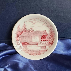 May include: A small, round, white dish with a red illustration of a covered bridge scene. The bridge is depicted with detailed lines, surrounded by trees and water. The dish has a slightly raised edge and is set against a blue background.