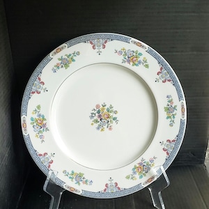 May include: White ceramic plate with a floral design. The plate features a blue Greek key border and floral clusters in the center and around the rim. The flowers are in shades of yellow, pink, and blue.