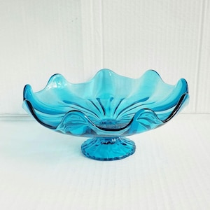 May include: A vintage, turquoise-colored glass pedestal bowl. The bowl has a scalloped rim and a round base. The glass is a vibrant shade of blue, and the bowl is likely used for serving fruit or displaying decorative items.