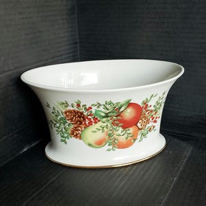 May include: White ceramic decorative bowl with a gold rim. The bowl features a colorful fruit and foliage design, including apples, pears, and pinecones. The bowl has a flared shape and a wide opening.
