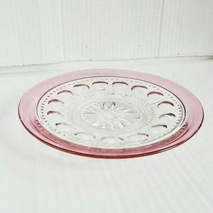 May include: A round glass plate featuring a clear center with a pressed starburst pattern, surrounded by a ring of embossed crescent shapes. The outer rim of the plate is tinted in a translucent pink hue, creating a distinct color contrast.