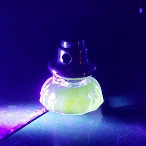 May include: A black metal bottle stopper with a rounded top and a hole, atop a glowing, light green, faceted glass base. The object is illuminated against a dark blue background.
