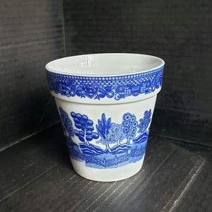 May include: A white ceramic flower pot with a blue Willow pattern design. The pot features a detailed landscape scene with trees, fences, and buildings. The rim has a decorative border. A classic piece for home decor.