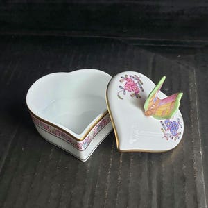 May include: A white heart-shaped porcelain trinket box with a matching lid. The lid features a three-dimensional butterfly figurine, pink floral patterns, and blue flowers. The base and lid edges are trimmed with gold and a thin decorative border.
