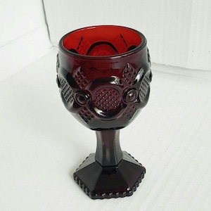 May include: A deep red glass goblet featuring a textured geometric pattern on the bowl. The vessel has a thick stem leading to a hexagonal base with a scalloped edge. The glass is translucent, showing a dark ruby hue throughout the entire piece.