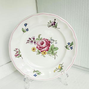May include: White ceramic plate with a scalloped edge and a pink rim. The plate features a central floral design with a large pink rose, green leaves, and smaller flowers in yellow, purple, and blue. Decorative tableware.