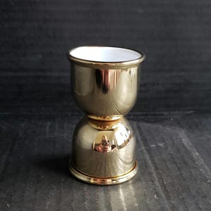 May include: A gold-colored double jigger with a white interior. The jigger has a rounded base and a wider top, with a gold rim on both ends. It is designed for measuring liquids, commonly used in bars.