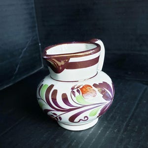May include: A white ceramic pitcher with a handle and a spout. The pitcher is decorated with brown stripes around the rim and floral patterns in purple, green, and orange. The pitcher is on a dark surface.