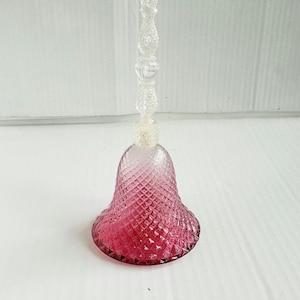 May include: A clear glass bell with a pink, diamond-patterned base and a clear, ornate handle. The bell is a decorative item, likely for display or collecting.