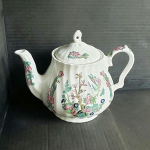 May include: White ceramic teapot with a floral design. The teapot features a curved spout, a handle, and a lid with a small knob. The floral pattern includes pink, green, and blue flowers and foliage. The teapot has a ribbed texture.