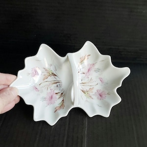 May include: A white ceramic divided serving dish shaped like two conjoined leaves. The dish features a delicate floral pattern with light pink petals and brown foliage accents, centered by a vertical handle with a thin gold trim along the edge.