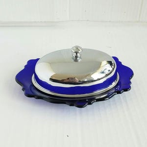 May include: A cobalt blue glass butter dish with a silver-colored metal lid. The dish has a scalloped edge and a small knob on top of the lid. The dish is designed to hold butter and keep it fresh.