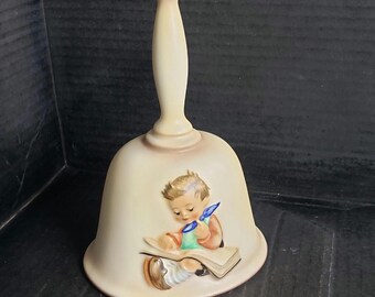 May include: A cream-colored ceramic bell with a long handle, featuring a hand-painted illustration of a young child with brown hair wearing a green shirt and orange sleeves, sitting while reading an open book with a blue quill pen in hand.