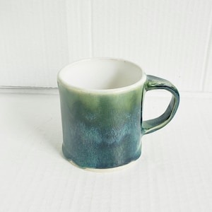 May include: A ceramic mug featuring a glossy glaze that transitions from a light green at the top to a deep teal and blue at the base. The mug has a smooth, white interior and a matching green handle, with a slightly wavy, uneven bottom edge.