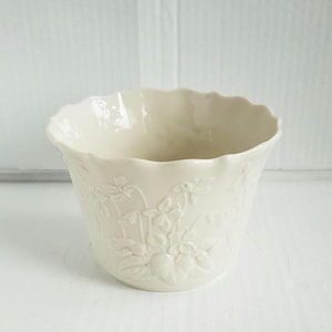 May include: Off-white ceramic planter with a scalloped rim and embossed floral design. The planter has a rounded shape and is suitable for small plants or decorative use. The embossed pattern features leaves and flowers.