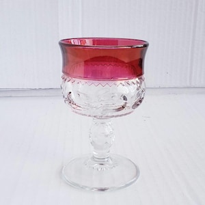 May include: A clear glass wine goblet with a ruby red rim and a decorative etched pattern on the bowl. The stem and base are also clear, creating a classic and elegant look. The goblet is designed for serving wine.