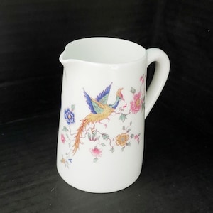 May include: White ceramic pitcher with a handle and a spout. The pitcher is decorated with a colorful bird and floral design. The bird has blue, yellow, and orange feathers. Pink, blue, and yellow flowers are also featured.