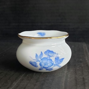 May include: A small, white ceramic bowl with a rounded shape and a gold rim. The bowl is decorated with blue floral designs. The bowl is likely used for decorative purposes or for holding small items.