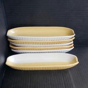 May include: A stack of six ceramic serving dishes. The dishes are oblong with a corn-on-the-cob design. The top half of each dish is yellow, and the bottom half is white. The dishes are approximately 10 inches long.