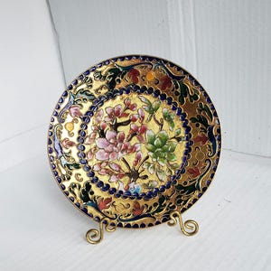May include: A decorative gold-colored plate with a floral design. The plate features a gold rim and a blue border with a floral pattern in pink, green, and red. The plate is displayed on a gold-colored metal stand.