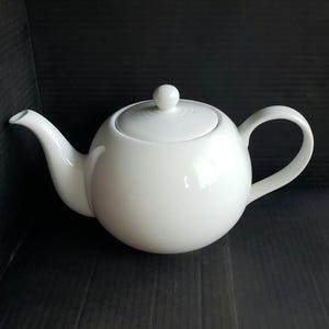 May include: A white ceramic teapot with a rounded body, a curved spout, and a loop handle. The lid has a small, round knob. The teapot is designed for brewing tea and has a classic, minimalist aesthetic.