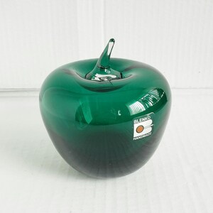 May include: A dark green glass apple sculpture with a clear glass stem. The apple features a white rectangular sticker on the front with the text BLENKO and HANDMADE printed in black, along with a circular orange graphic icon.