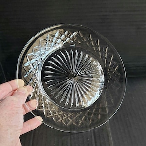 May include: A clear glass plate featuring a central sunburst pattern of radiating lines surrounded by a wide border of pressed diamond-shaped facets. The circular dish has a smooth, transparent finish and a distinct geometric texture.