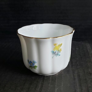 May include: White porcelain teacup with a scalloped rim and a gold-colored band. The cup features delicate floral designs in yellow and blue. The cup has a fluted body and is set against a dark background.