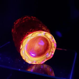 May include: A glowing, cylindrical glass object with a textured exterior and a wide, open top. The interior glows with a bright yellow and orange hue, with a purple center. The object is set against a dark background.