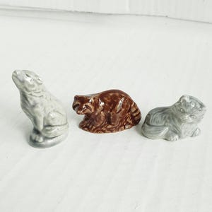 May include: Three ceramic animal figurines: a gray wolf, a brown raccoon, and a gray cat. The wolf is standing, the raccoon is crouched, and the cat is lying down. Each figurine has detailed features and a glossy finish.
