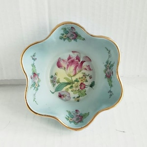 May include: A light blue, scalloped-edge dish with a gold rim. The center features a pink and white tulip with yellow and green accents. Floral sprigs decorate the dish's sides, creating a vintage aesthetic.