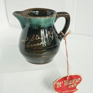 May include: A small, handcrafted ceramic pitcher with a dark brown base and a teal green rim. The pitcher has a handle and a spout. The words "Sault Ste. Marie Canada" are written on the side. A tag reads "McMaster craft, Dundas, Canada."