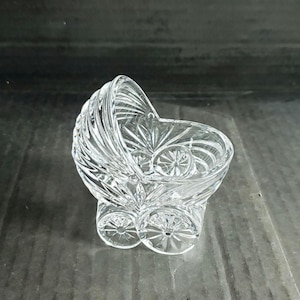 May include: A clear glass figurine shaped like a vintage baby carriage, featuring intricate cut patterns on the hood, body, and wheels. The glass has a reflective, faceted surface that catches light, creating a detailed, decorative miniature object.