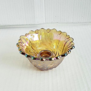 May include: A decorative, iridescent glass bowl with a ruffled edge. The bowl has a golden-yellow hue with hints of blue and purple. It features a small, circular indentation in the center, likely for a candle or small item.