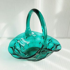 May include: A teal-colored glass basket with a handle. The basket has a diamond pattern and a scalloped edge. The handle arches gracefully over the bowl. The glass is transparent, allowing light to pass through, and the basket is likely used for decorative purposes.