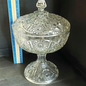 May include: A clear pressed glass compote dish with a lid, featuring a faceted finial and intricate starburst and crosshatch patterns. A wooden ruler placed vertically next to the dish indicates a total height of approximately 9.5 inches.
