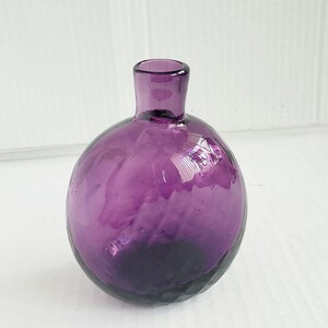 May include: A small, spherical purple glass vase with a narrow, cylindrical neck. The glass features a subtle, dimpled, or hammered texture across its rounded body, creating light reflections on the translucent, deep violet surface.
