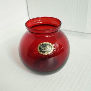 May include: A round, deep red glass vase with a wide, open mouth. A gold and white oval sticker is affixed to the front, featuring the text Anchor Hocking Glass Corp, Royal Ruby, and Lancaster, Ohio, USA, in a clear, legible font style.