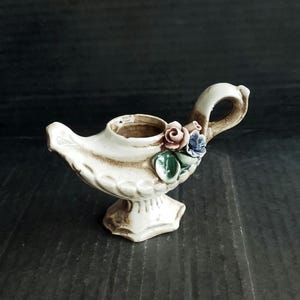 May include: A white ceramic genie lamp-shaped vase with a handle and a spout. The vase is decorated with a cluster of pink, blue, and green floral accents. The base is pedestal-style.
