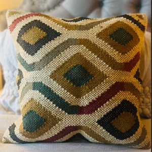 May include: A square throw pillow with a geometric pattern in shades of brown, green, red, and black. The pillow is made of a woven fabric with a textured surface.