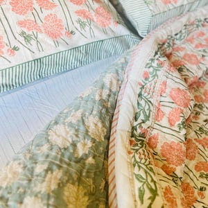 May include: A white and green floral patterned quilt with a pink floral design. The quilt is folded over a white and blue striped sheet.