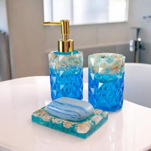 Könnte beinhalten: Ein blaues und weißes Badezimmer-Set mit einem Seifenspender, einem Becher und einem Seifenschälchen. Das Set ist mit Muscheln verziert und hat ein Strandthema.